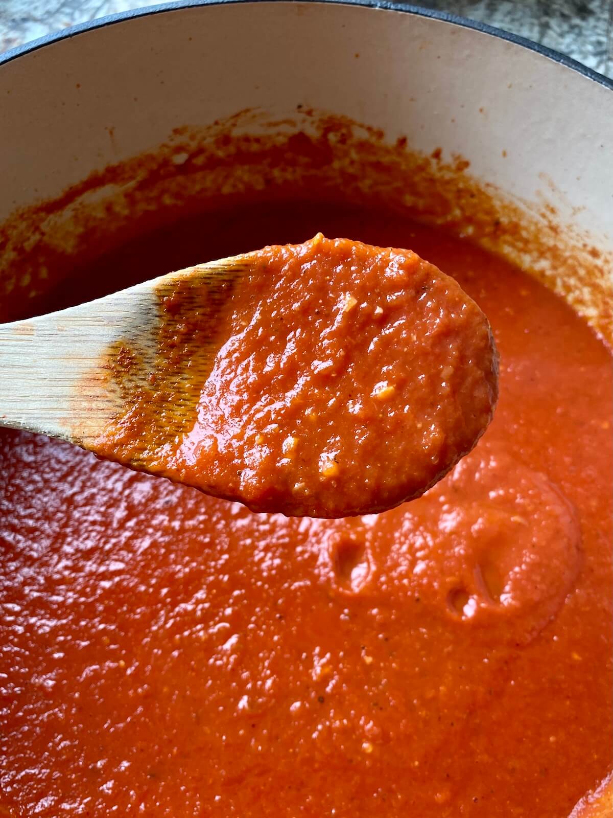 A wooden spoon holding up a scoop of tomato sauce over a Dutch oven filled with the rest of the tomato sauce.