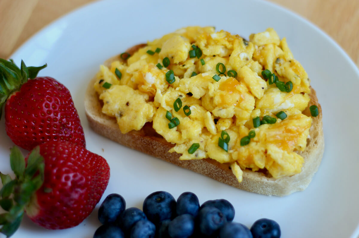 Scrambled eggs on toast on a small white plate. Next to the piece of toast with eggs is two strawberries and a small pile of blueberries.