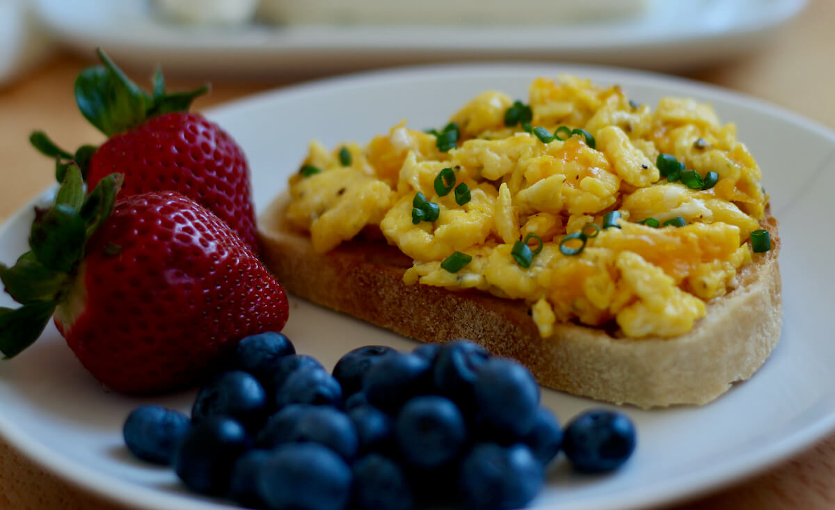 Scrambled eggs on toast next to two strawberries and some blueberries on a small white plate. Out of focus in the background is a stick of butter on a butter dish.