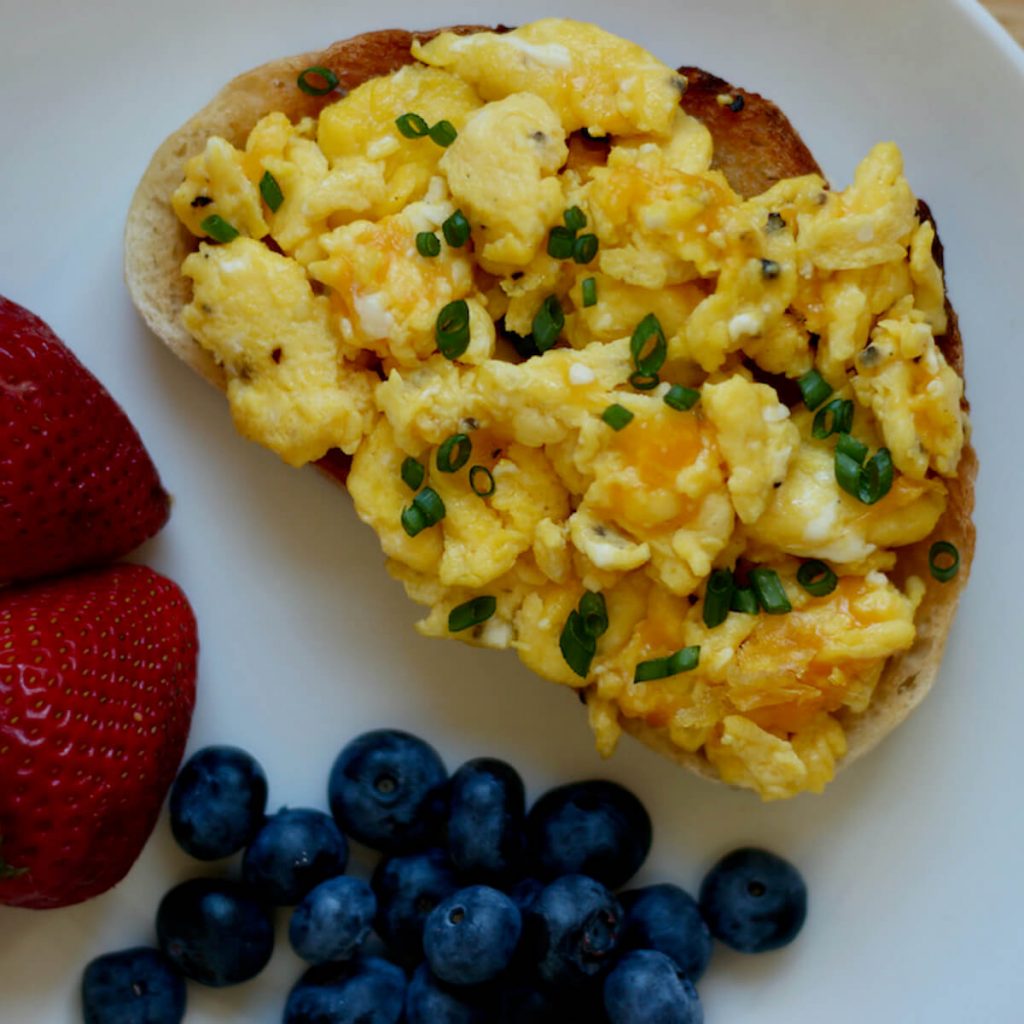 A slice of toasted sourdough bread topped with scrambled eggs, cheddar cheese, and chives on a white plate. To the side is two strawberries and a handful of blueberries.