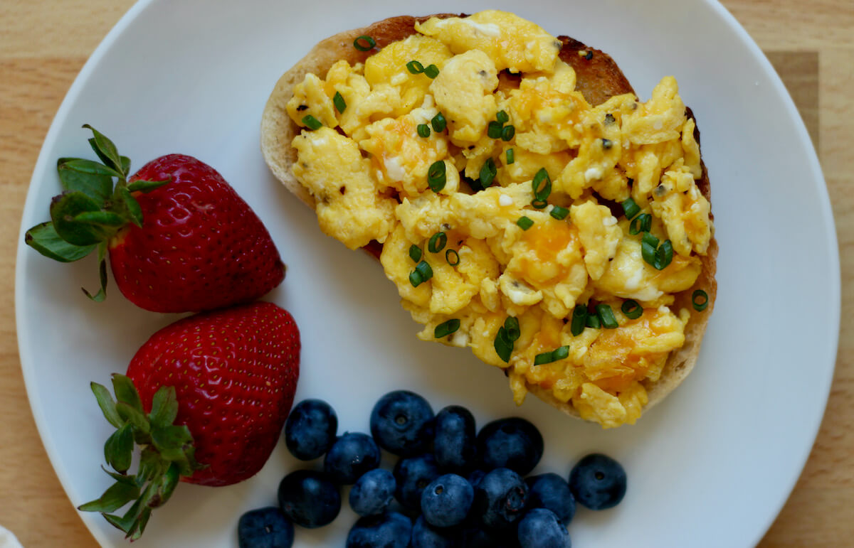 A small, white plate with a slice of bread with scrambled eggs on top. Beside the toast is two strawberries and a small pile of blueberries.