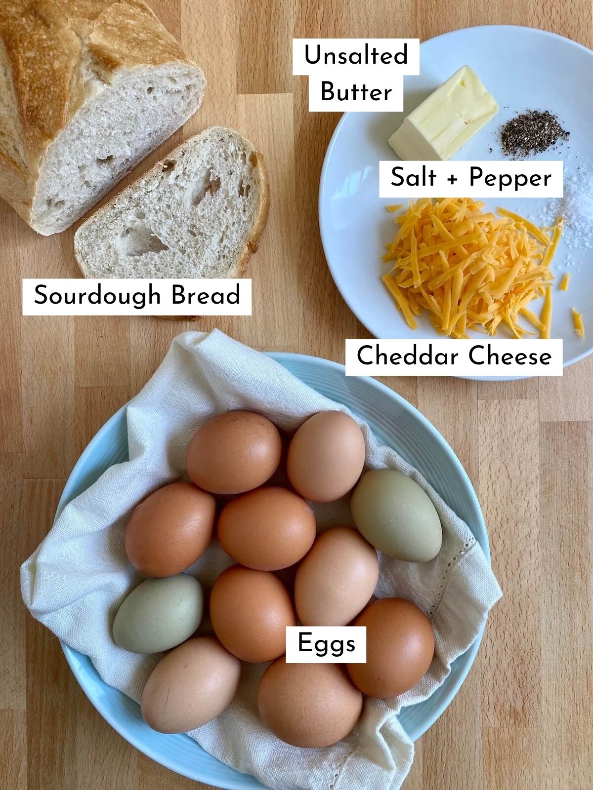 The ingredients to make scrambled eggs on toast on a butcher block countertop. There is text over each ingredient, stating what it is. The ingredients include sourdough bread, unsalted butter, eggs, cheddar cheese, salt, and pepper.