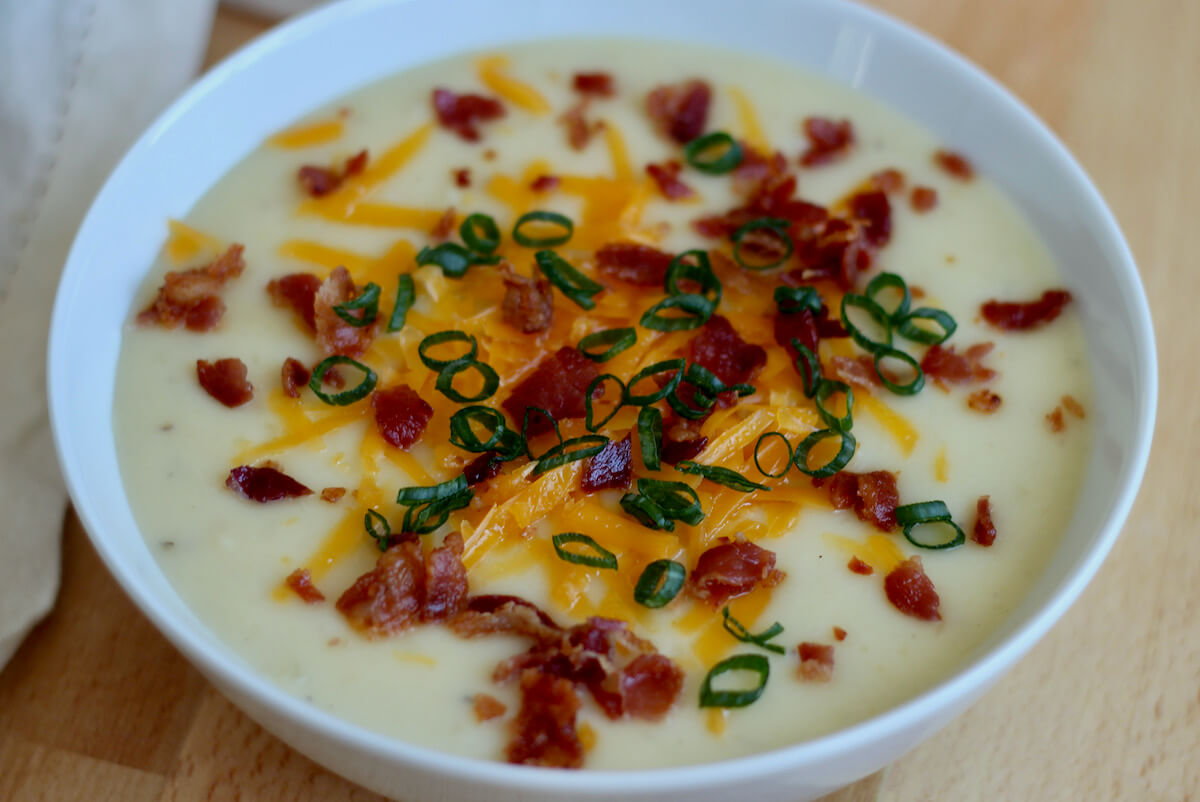 A bowl of potato soup topped with cheddar cheese, bacon, and green onion on a countertop.