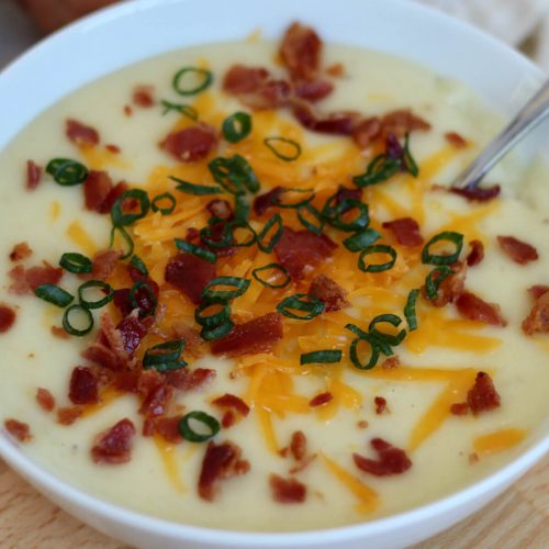 A white bowl filled with 4 ingredient potato soup. The soup is topped with shredded cheddar cheese, crumbled bacon, and scallions. There is a silver spoon sticking out of the soup bowl to the right.