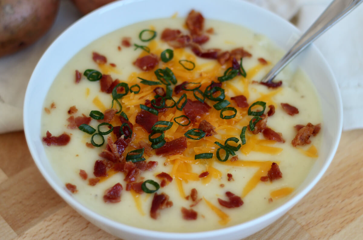 A white bowl filled with 4 ingredient potato soup, which is topped with shredded cheddar cheese, crumbled bacon bits, and sliced green onion. There are two potatoes out of focus in the background.