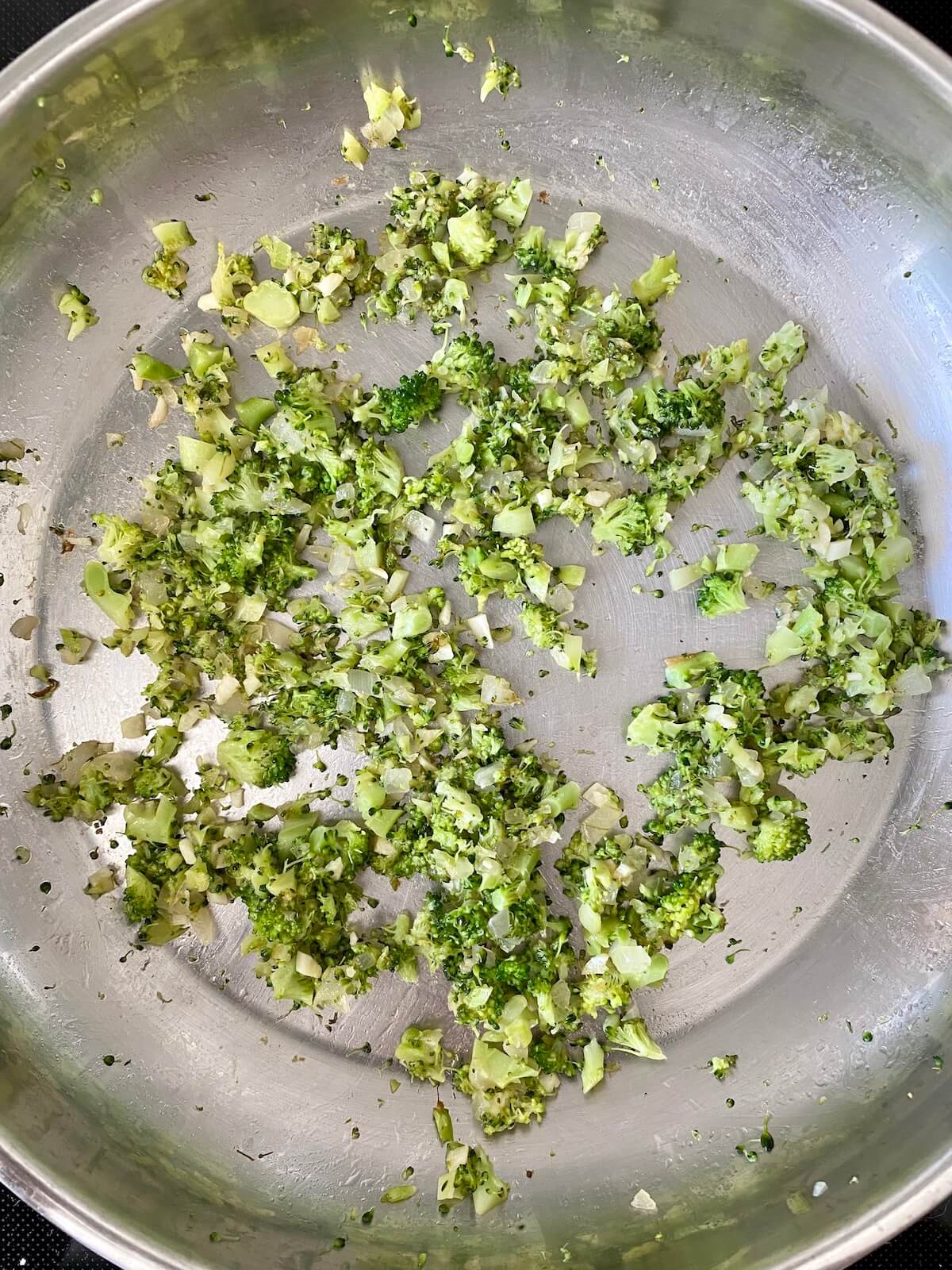 Finely chopped broccoli florets, onion, and garlic cooking in a small stainless steel pan.