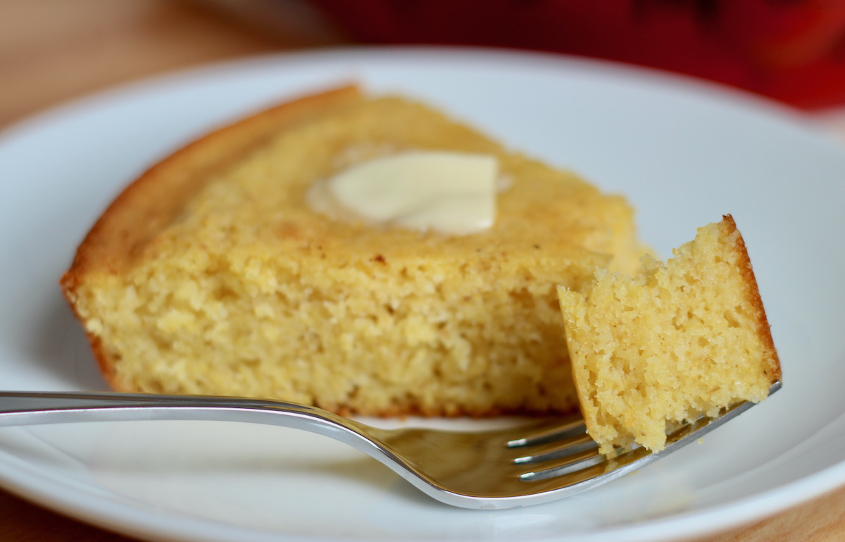 A fork holding a bite of skillet cornbread on a small white plate. Out of focus on the plate is the remaining slice of cornbread with a pat of butter melting on top of it.