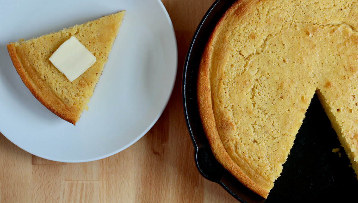 Cornbread in a cast iron skillet with a single slice removed. Next to the cast iron skillet is a small white place with a slice of cornbread on it. There is a small pat of butter on top of the slice of cornbread.
