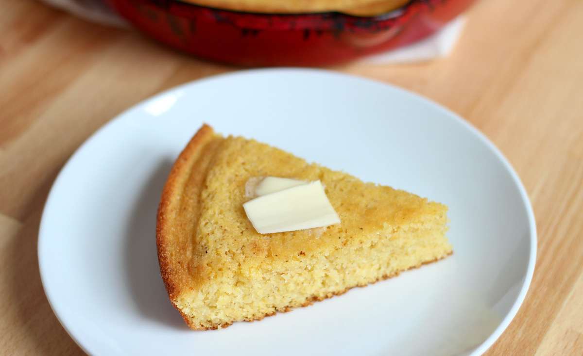 A slice of brown butter cornbread with a pat of butter on top of it is sitting on a small white plate. The red cast iron skillet with the remaining cornbread is out of focus in the background.