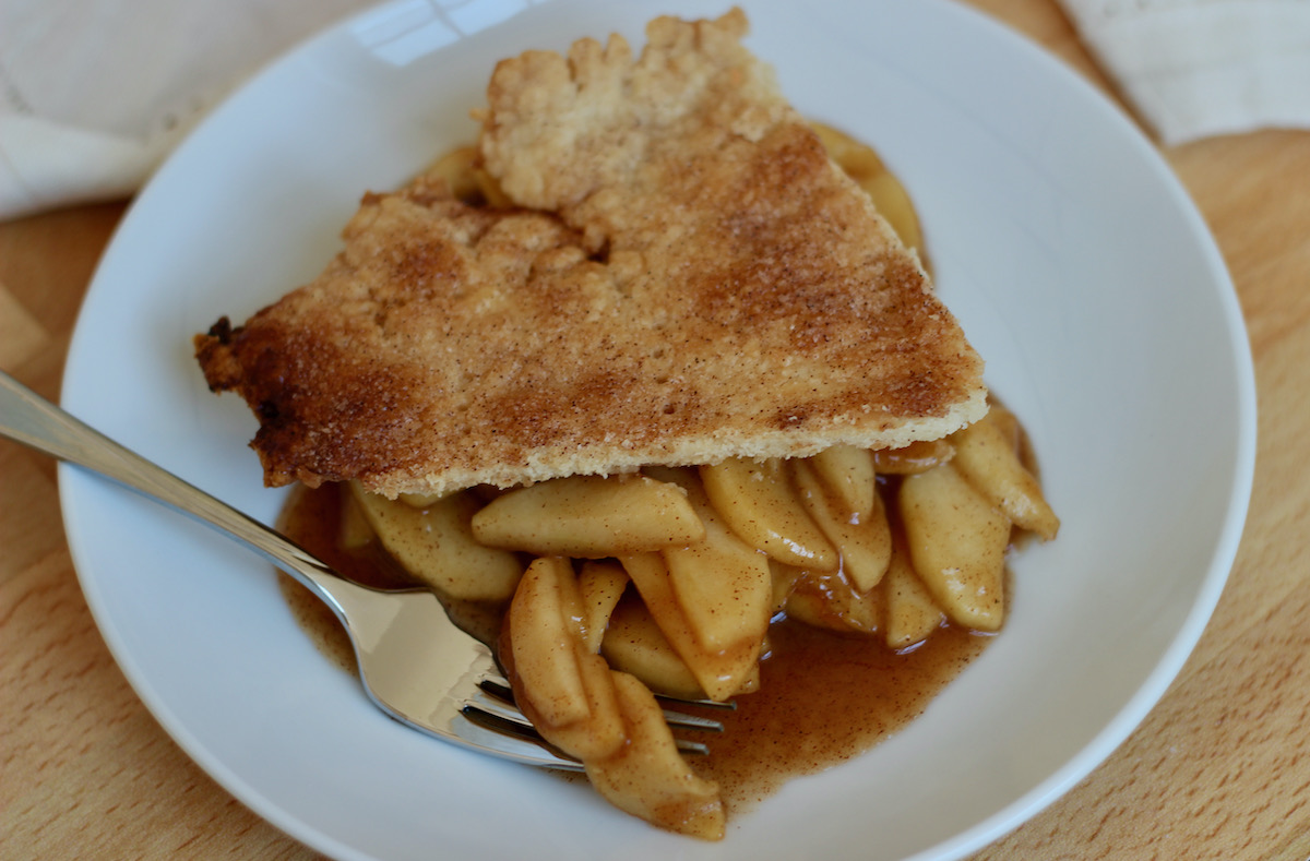 A serving of deconstructed apple pie on a small white plate. There is a fork on the plate with a few pieces of apple on it, as if someone is about to take a bite. There is a white cloth napkin out of focus in the background.
