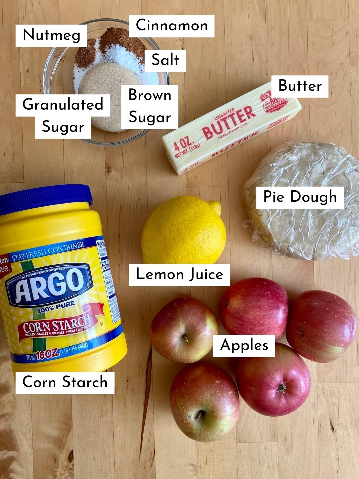 The ingredients to make deconstructed apple pie displayed on a butcher block countertop. The ingredients are labeled with text stating what each ingredient is. The ingredients include apples, pie dough, butter, brown sugar, granulated sugar, cinnamon, nutmeg, salt, lemon juice, and corn starch.