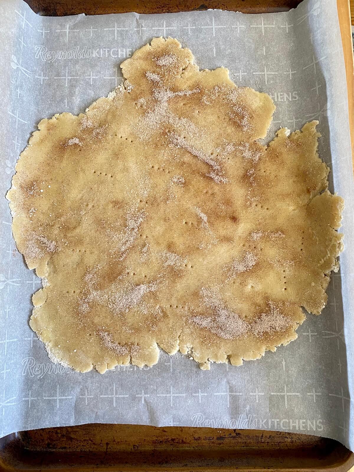 A rolled out pie dough on a sheet of parchment paper on a baking sheet. There is cinnamon and sugar sprinkled liberally on the pie dough.