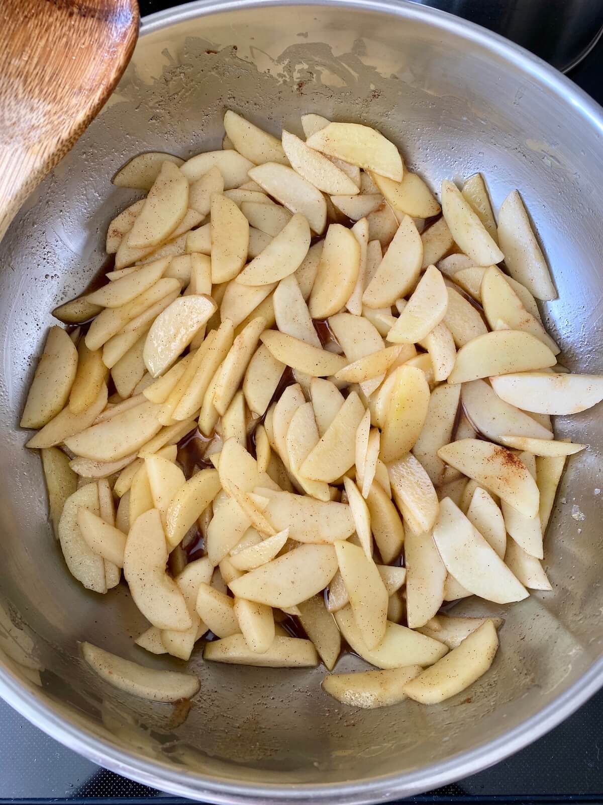 Thinly sliced apples being sautéed with cinnamon, sugar, nutmeg, and butter in a stainless steel skillet. There is a wooden spoon sitting on the left edge of the pan.