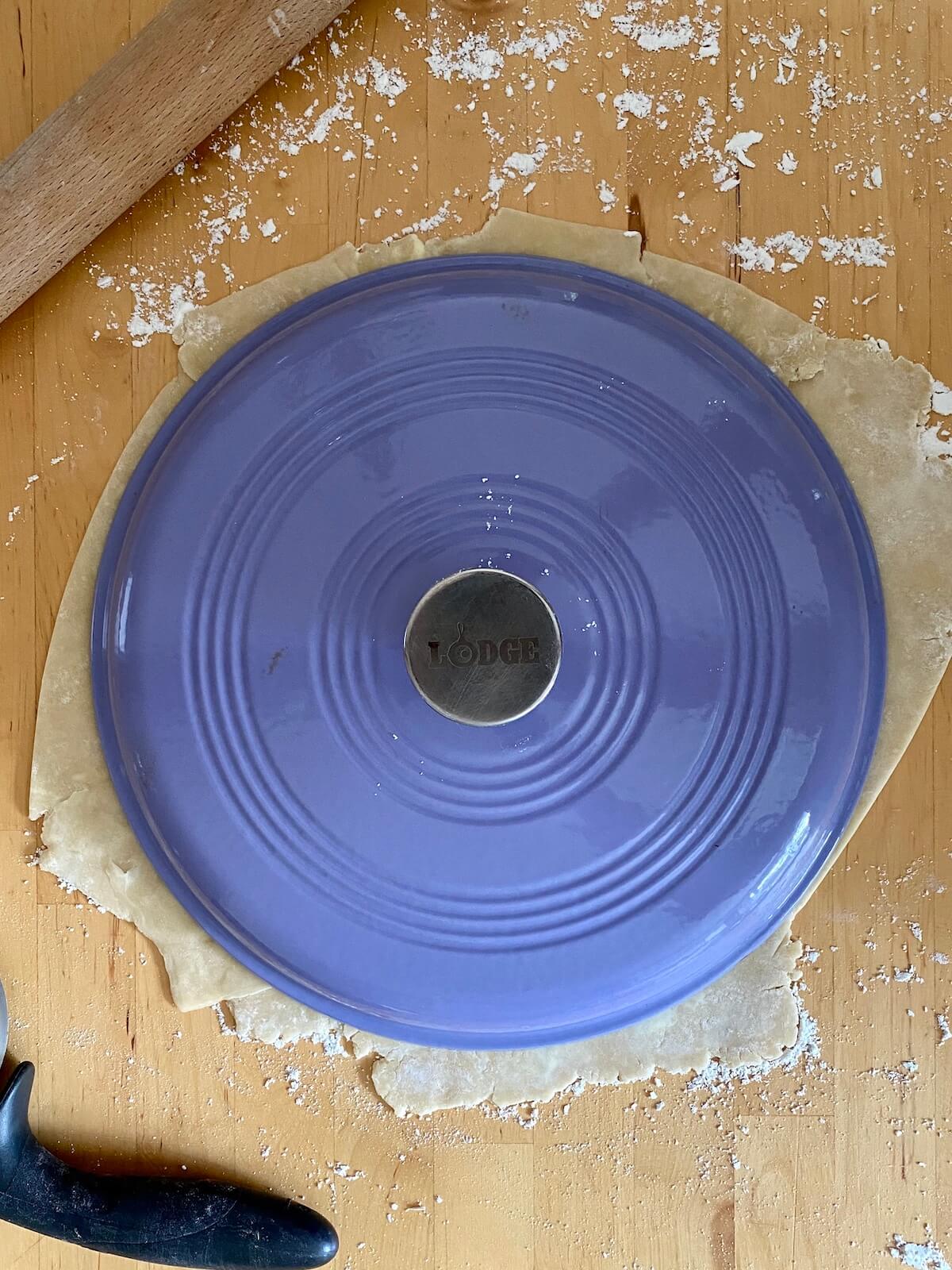 A rolled out pie dough with a purple dutch oven lid placed on top to measure it. There is flour sprinkled around the pie dough and the rolling pin and pizza cutter are off to the side.
