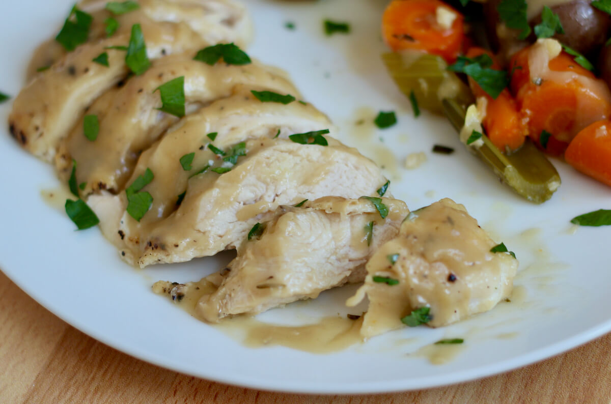 Sliced chicken breast on a white plate topped with homemade chicken gravy. The chicken is garnished with fresh parsley. Out of focus in the background are braised carrots, celery, and potatoes.
