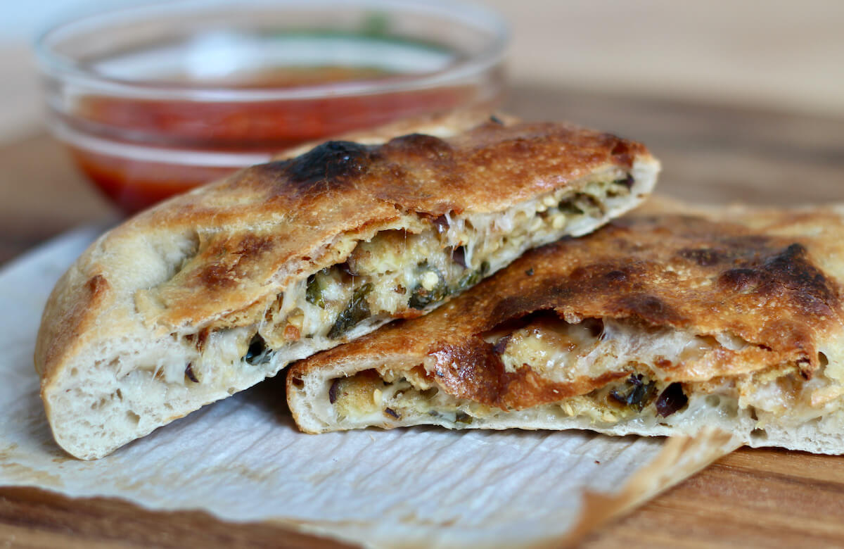 An eggplant calzone cut in half on a piece of parchment paper. There is a small glass bowl of marinara dipping sauce in the background.