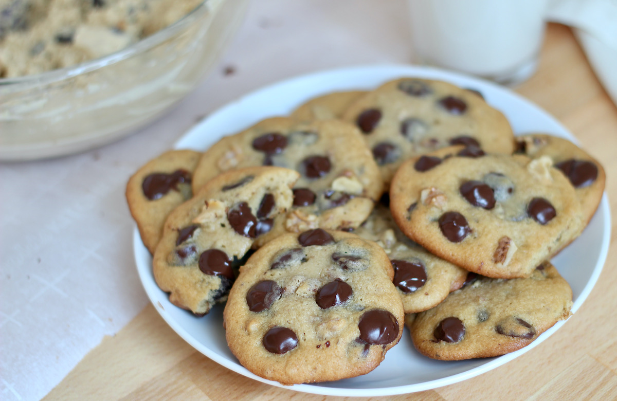 A small white plate piled with maple syrup chocolate chip cookies. Out of focus in the background is a glass of milk and the mixing bowl filled with the remaining cookie dough batter.