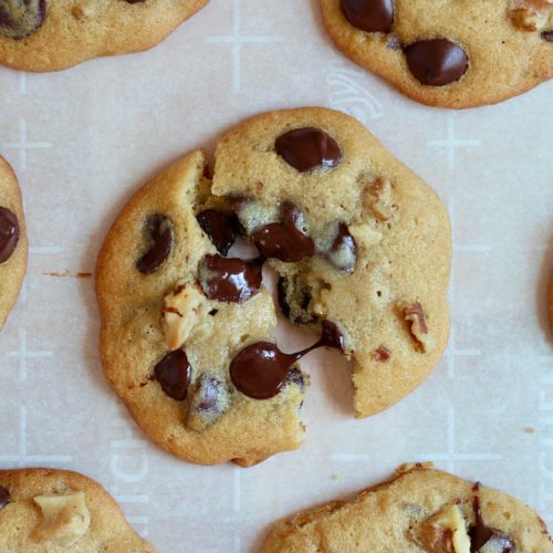 A maple syrup chocolate chip cookie on a sheet of white parchment paper. The cookie is split in half and the melted chocolate chips are still connecting the halves by a string of melted chocolate. There are other cookies around the halved cookie.