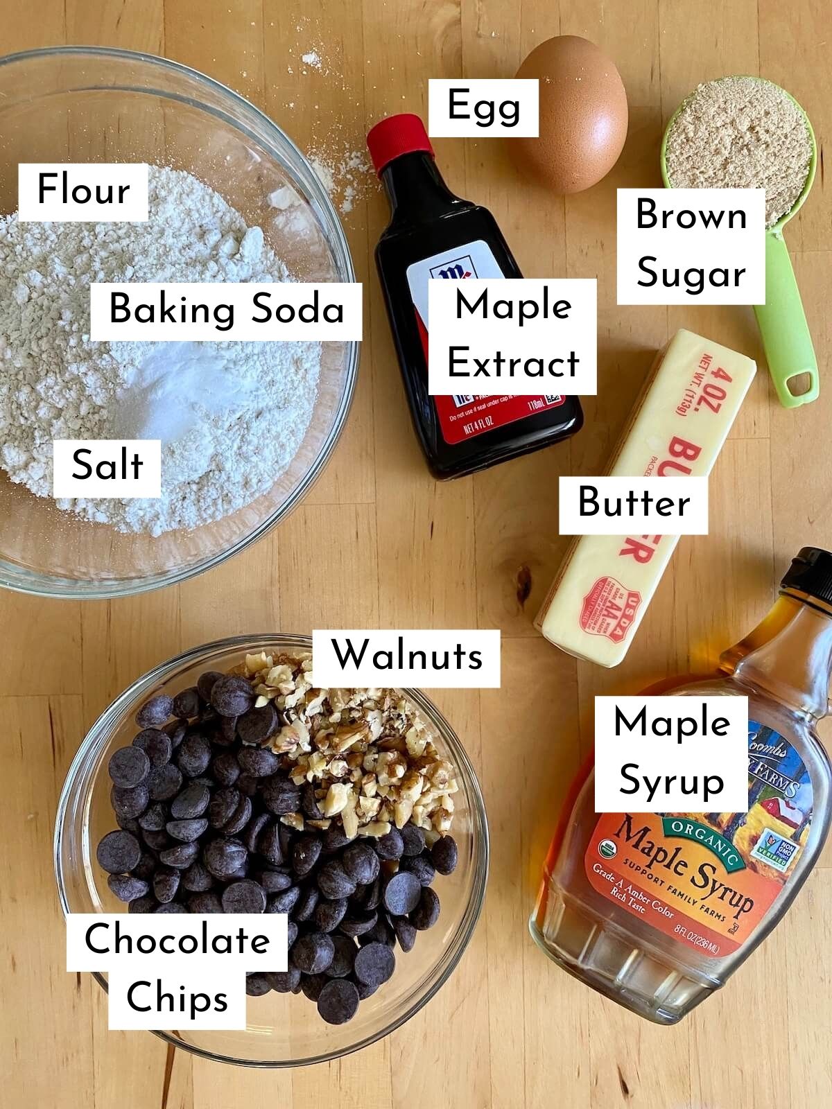 The ingredients to make maple syrup chocolate chip cookies on a butcher block countertop. There is text over each ingredient, stating what it is. The ingredients include flour, baking soda, salt, egg, brown sugar, maple extract, butter, maple syrup, walnuts, and chocolate chips.