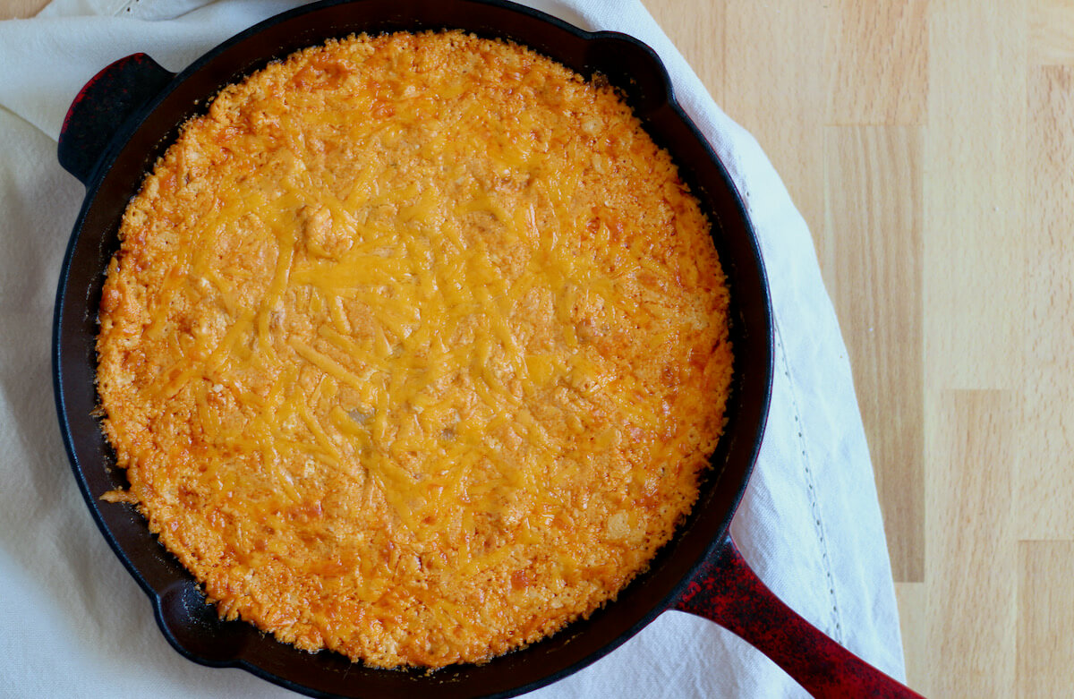 A cast iron skillet filled with cheesy, bubbly white bean buffalo dip. The cast iron skillet is sitting on a white cloth napkin on a butcher block countertop.