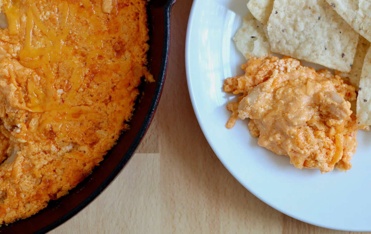 On the left is a cast iron skillet filled with white bean buffalo dip. On the right is a small white plate with a scoop of dip and some tortilla chips on it.