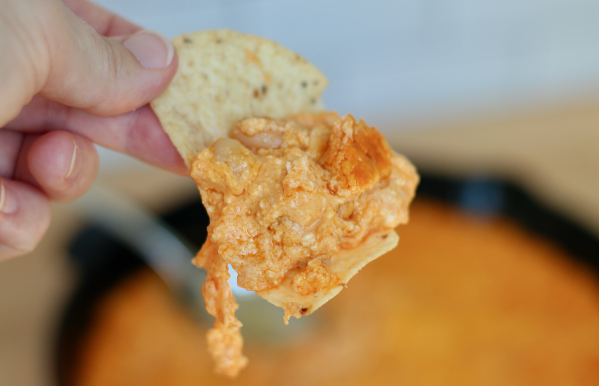 A hand holding a tortilla chip with white bean buffalo dip on it. Out of focus in the background is a cast iron skillet filled with the remaining dip.