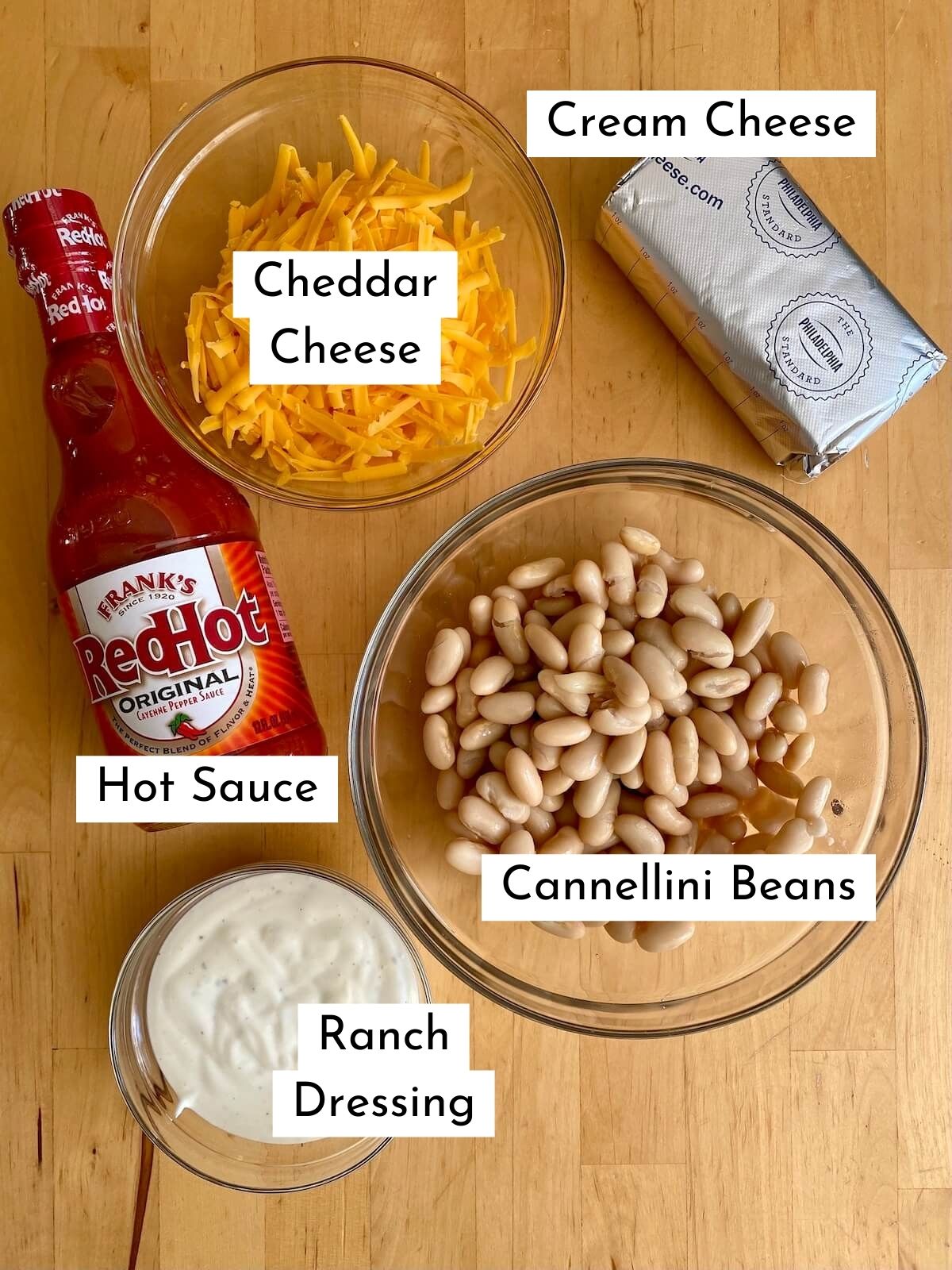 The ingredients to make buffalo white bean dip on a butcher block countertop. The ingredients have text over them that state what each ingredient is. The ingredients include cannellini beans, cream cheese, cheddar cheese, hot sauce, and ranch dressing.