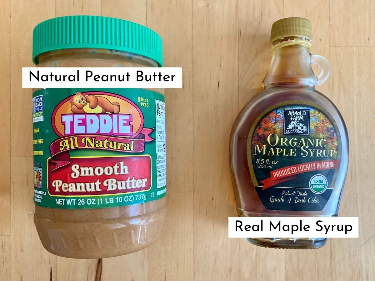 A jar of natural peanut butter and a bottle of real maple syrup on a butcher block countertop. The ingredients are labeled with text.