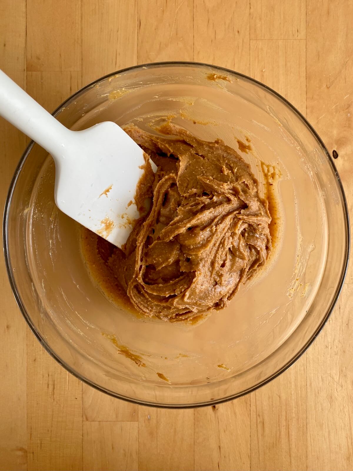 A clear glass bowl with peanut butter and maple syrup mixed together. There is a rubber spatula sticking out of the bowl to the left.