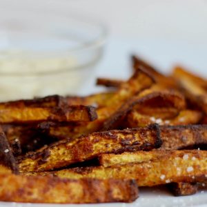A pile of crispy air fryer butternut squash fries garnished with kosher salt on a white plate. Out of focus in the background is a small glass bowl filled with a garlic aioli dipping sauce.