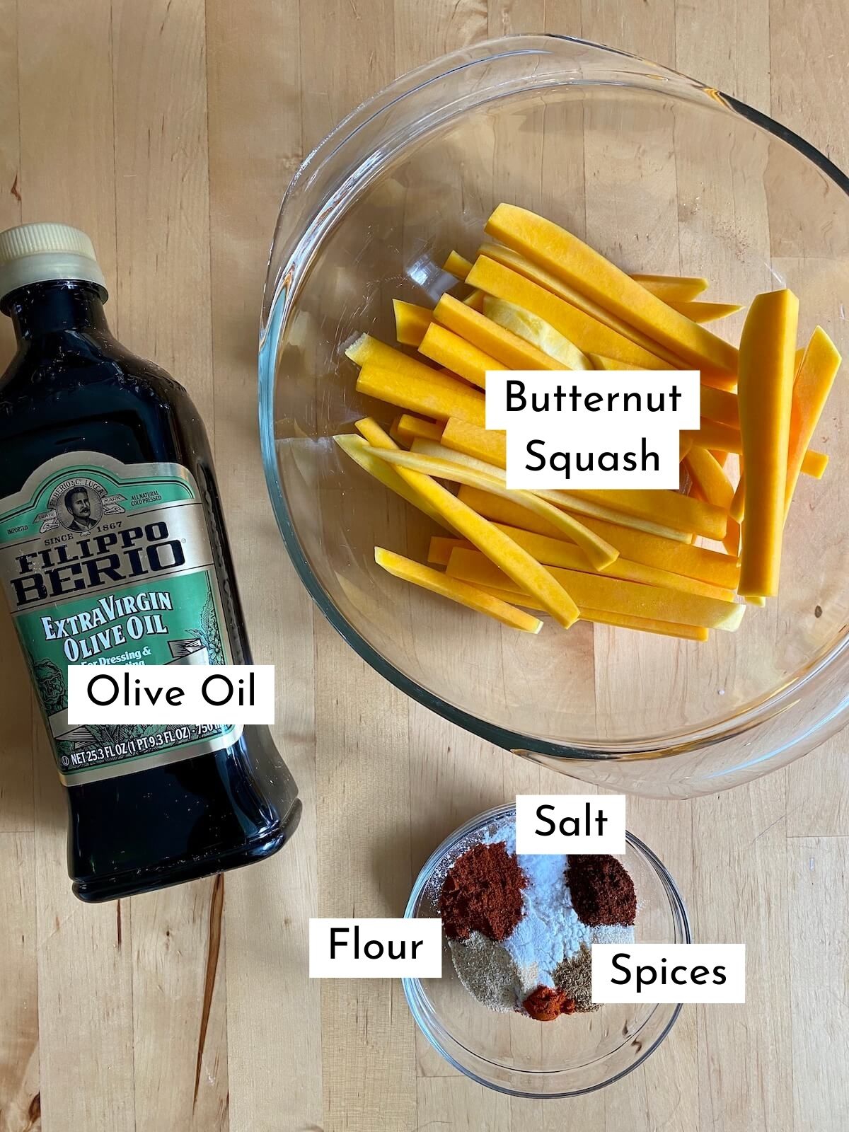 The ingredients to make air fryer butternut squash fries on a wooden counter. There is text over each ingredients, labeling what it is. The ingredients include olive oil, butternut squash, flour, salt, and spices.