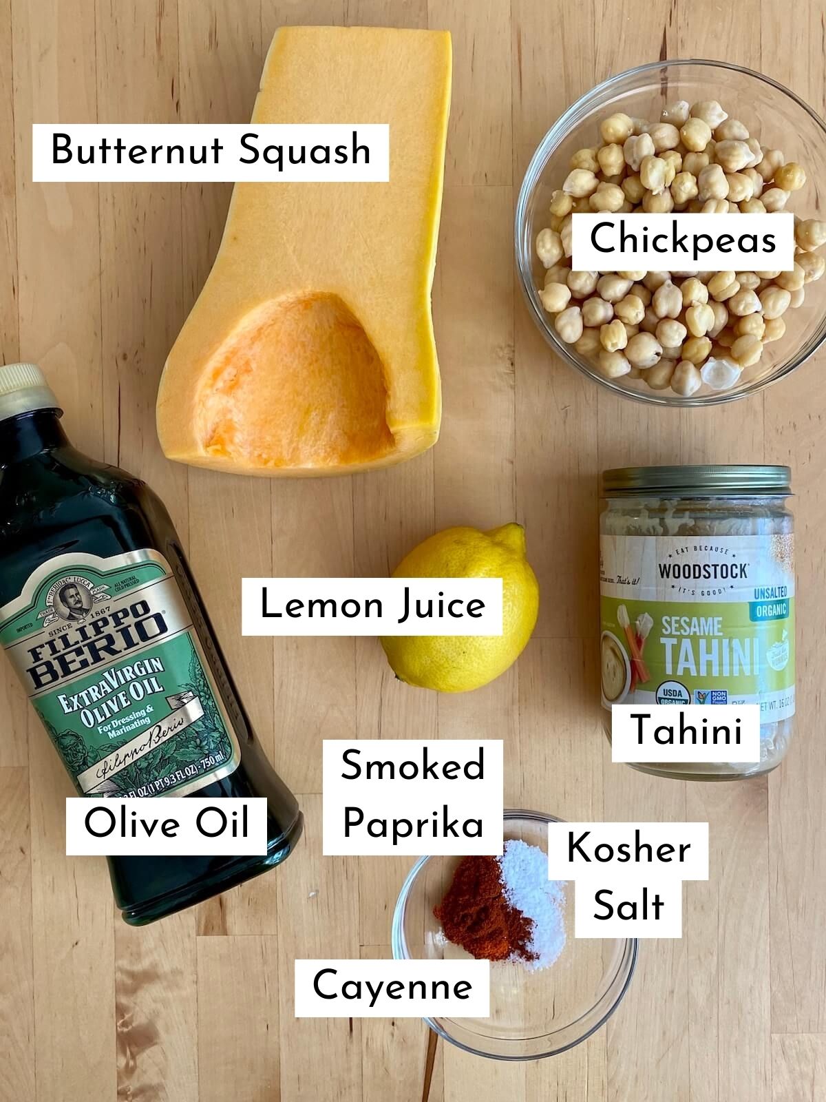 The ingredients to make butternut squash hummus on a wooden countertop. Each ingredient is labeled with text describing what it is. The ingredients include butternut squash, chickpeas, olive oil, lemon juice, tahini, smoked paprika, kosher salt, and cayenne.