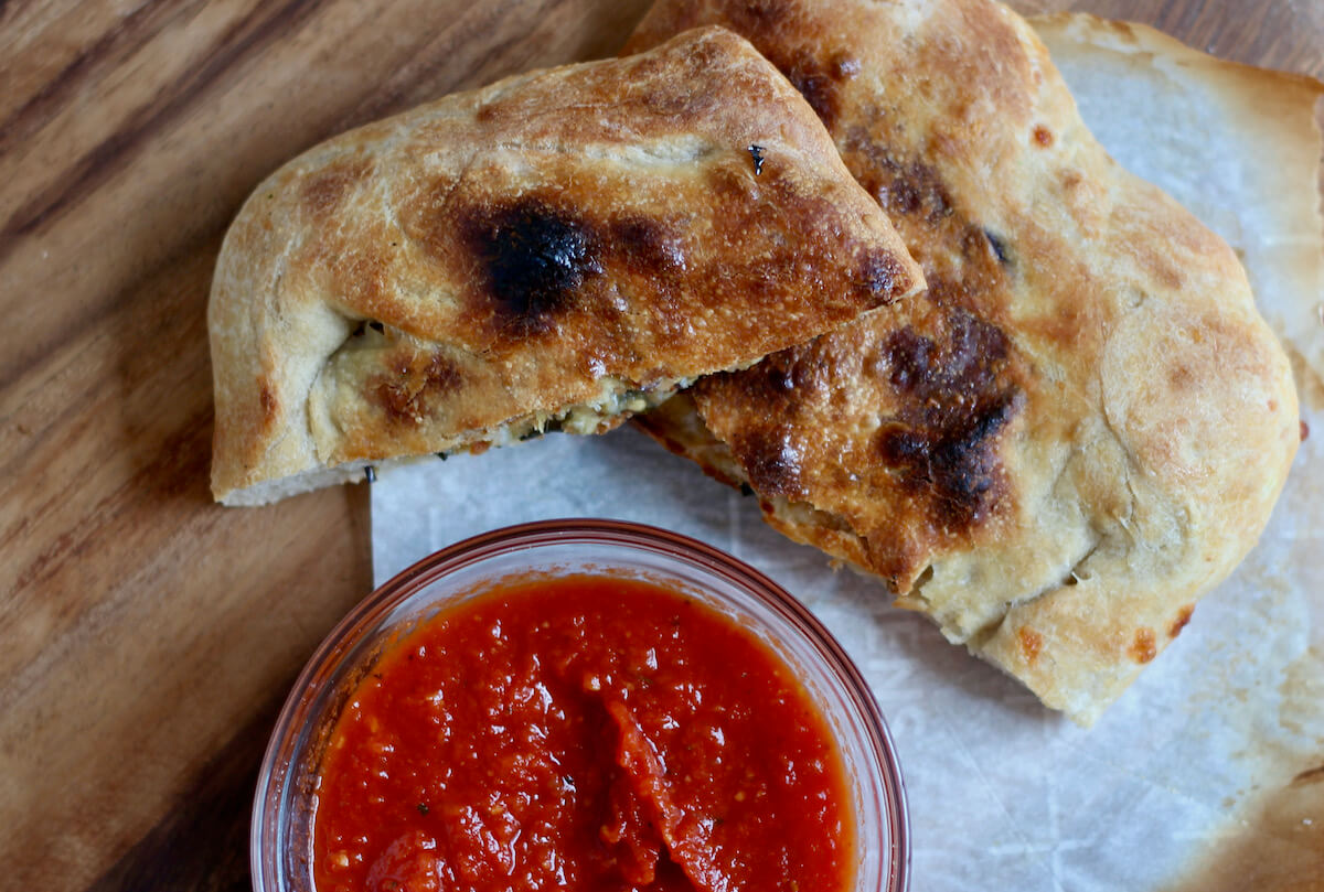 Two halves of an eggplant calzone stacked on top of one another next to a bowl of marinara sauce.