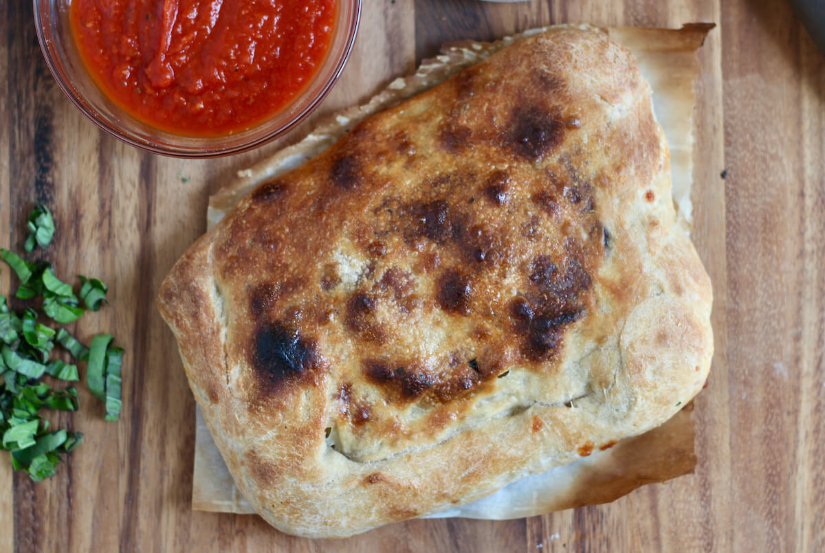 A whole eggplant parmesan calzone on a piece of parchment paper. Next to the calzone is some chopped fresh basil and a small bowl of marinara sauce.