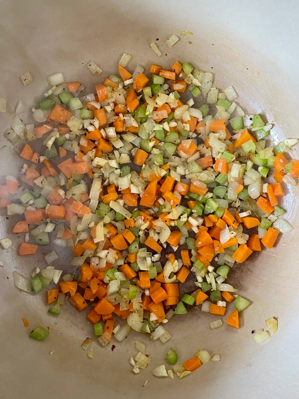 Onion, carrot, celery, garlic, salt, pepper, thyme, and a bay leaf being sautéed in olive oil in a Dutch oven.