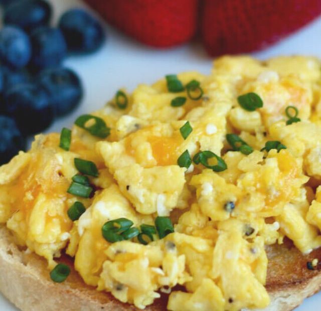 Scrambled eggs with cheddar cheese and chives on a piece of sourdough toast. Out of focus in the background is some strawberries and blueberries.