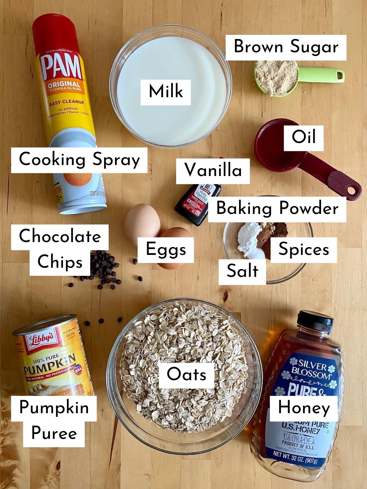 The ingredients to make pumpkin baked oats on a wooden countertop. There is text on top of each ingredient stating what it is. The ingredients include: cooking spray, milk, brown sugar, oil, vanilla, baking powder, salt, spices, eggs, chocolate chips, pumpkin puree, oats, honey.