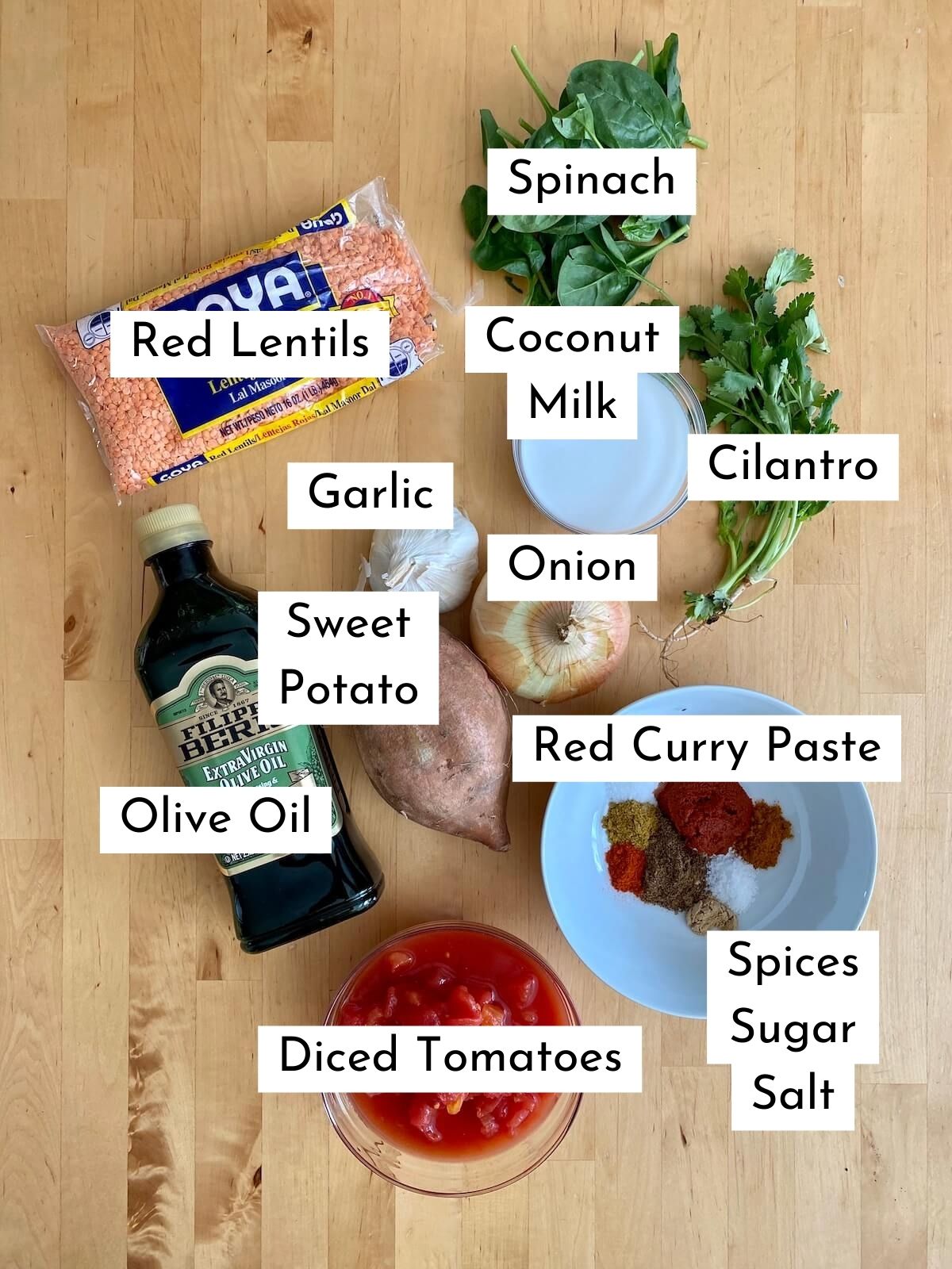 The ingredients to make red lentil and sweet potato curry laid out on a butcher block countertop. Each ingredient is labeled with text. The ingredients include red lentils, spinach, garlic, onion, sweet potato, coconut milk, olive oil, diced tomatoes, red curry paste, cilantro, spices, sugar, and salt.