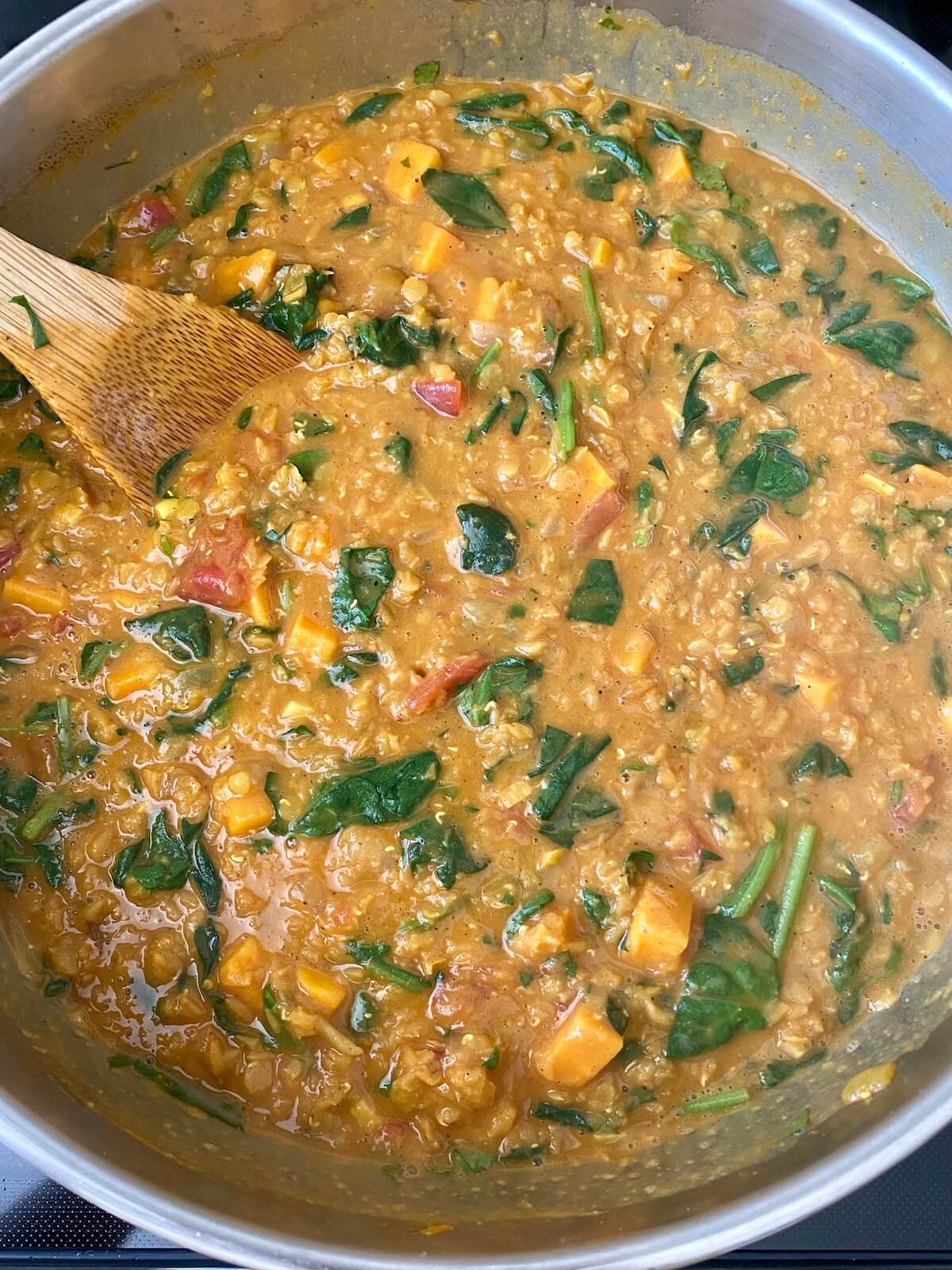 Finished red curry lentils with sweet potatoes and spinach in a stainless steel skillet. There is a wooden spoon sticking out of the pot to the left.