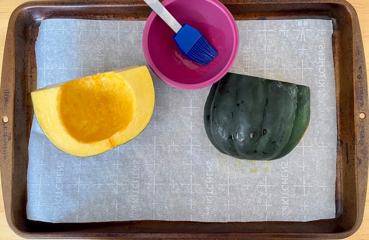 Raw, halved acorn squash on a parchment-lined baking sheet after being brushed with olive oil.