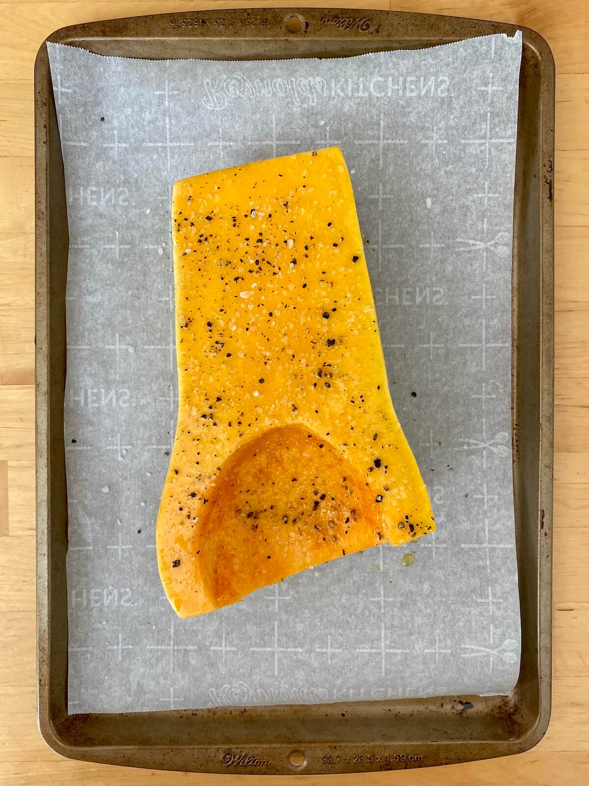 A half of a butternut squash seasoned with salt, pepper, and olive oil on a parchment lined baking sheet before being roasted.