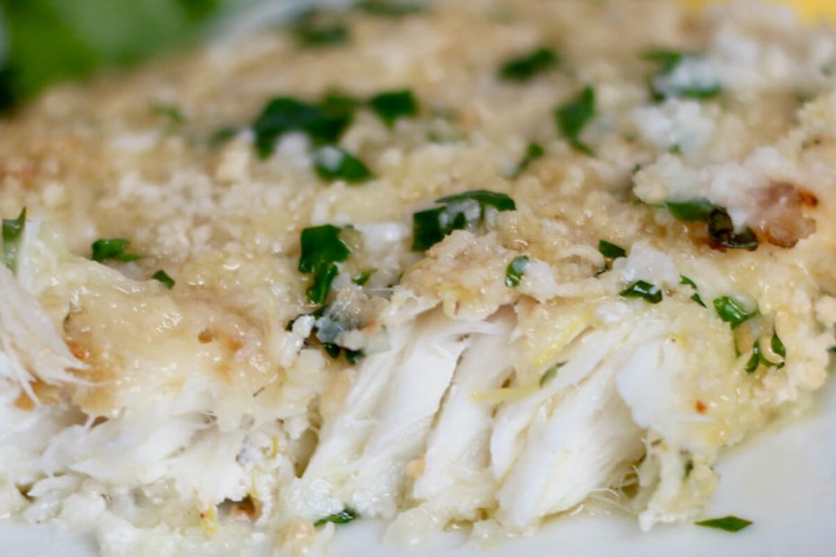 A piece of parmesan crusted air fryer halibut on a small white plate. The fish is garnished with butter and parsley and a piece has been removed so you can see the white flaky fish.