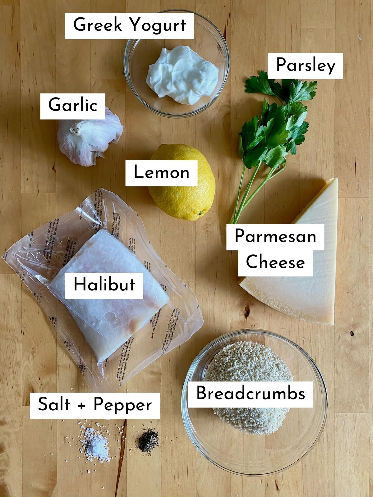 The ingredients to make air fryer halibut on a wooden countertop. The ingredients are labeled with text and they include Greek yogurt, garlic, lemon, parsley, halibut, parmesan cheese, breadcrumbs, salt, and pepper.