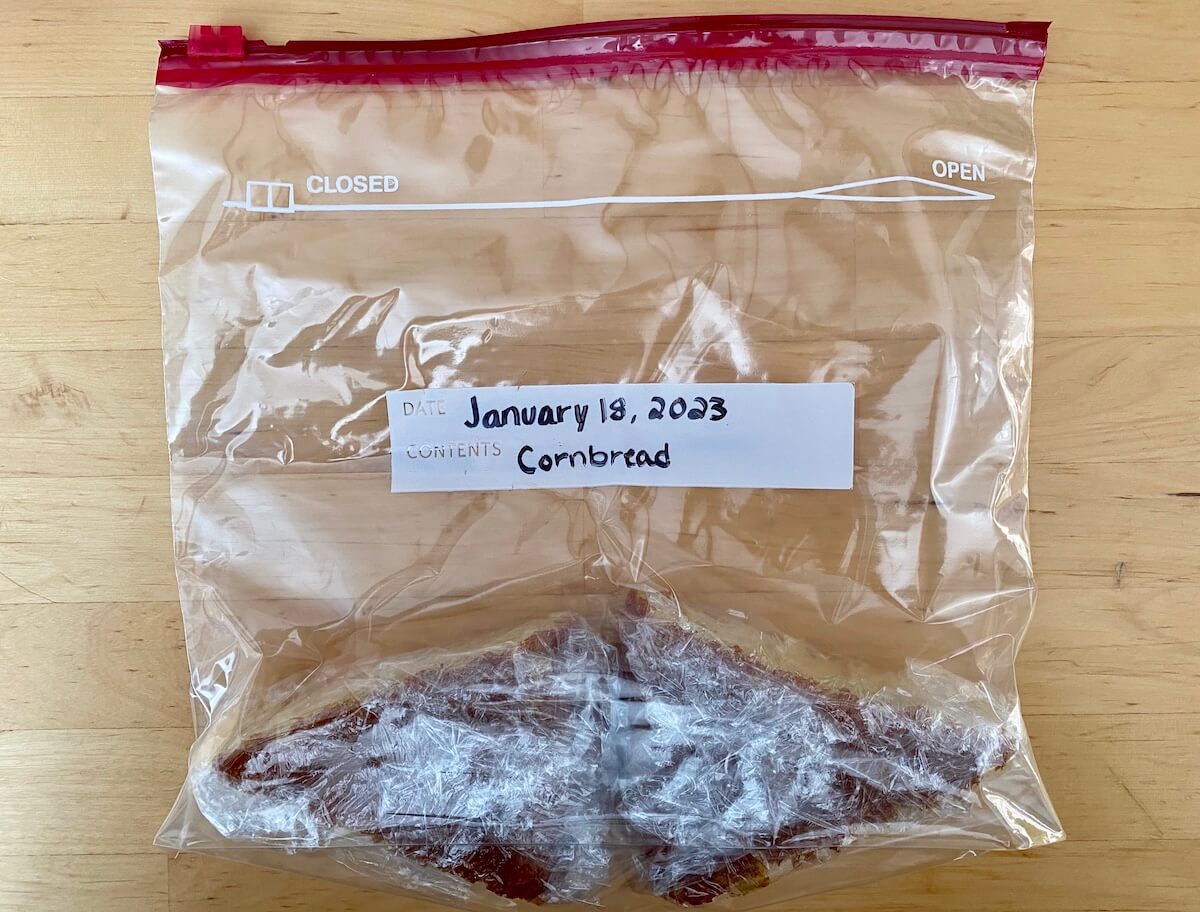 Two pieces of cornbread wrapped in plastic wrap inside of a freezer-safe plastic zipper bag. The bag is dated January 18, 2023 and has the word "cornbread" written on it.