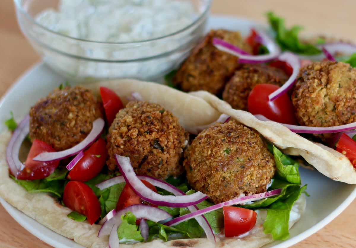 Two homemade falafel gyros with lettuce, red onion, and tomatoes on a small white plate. There is a small bowl of tzatziki on the plate in the background.