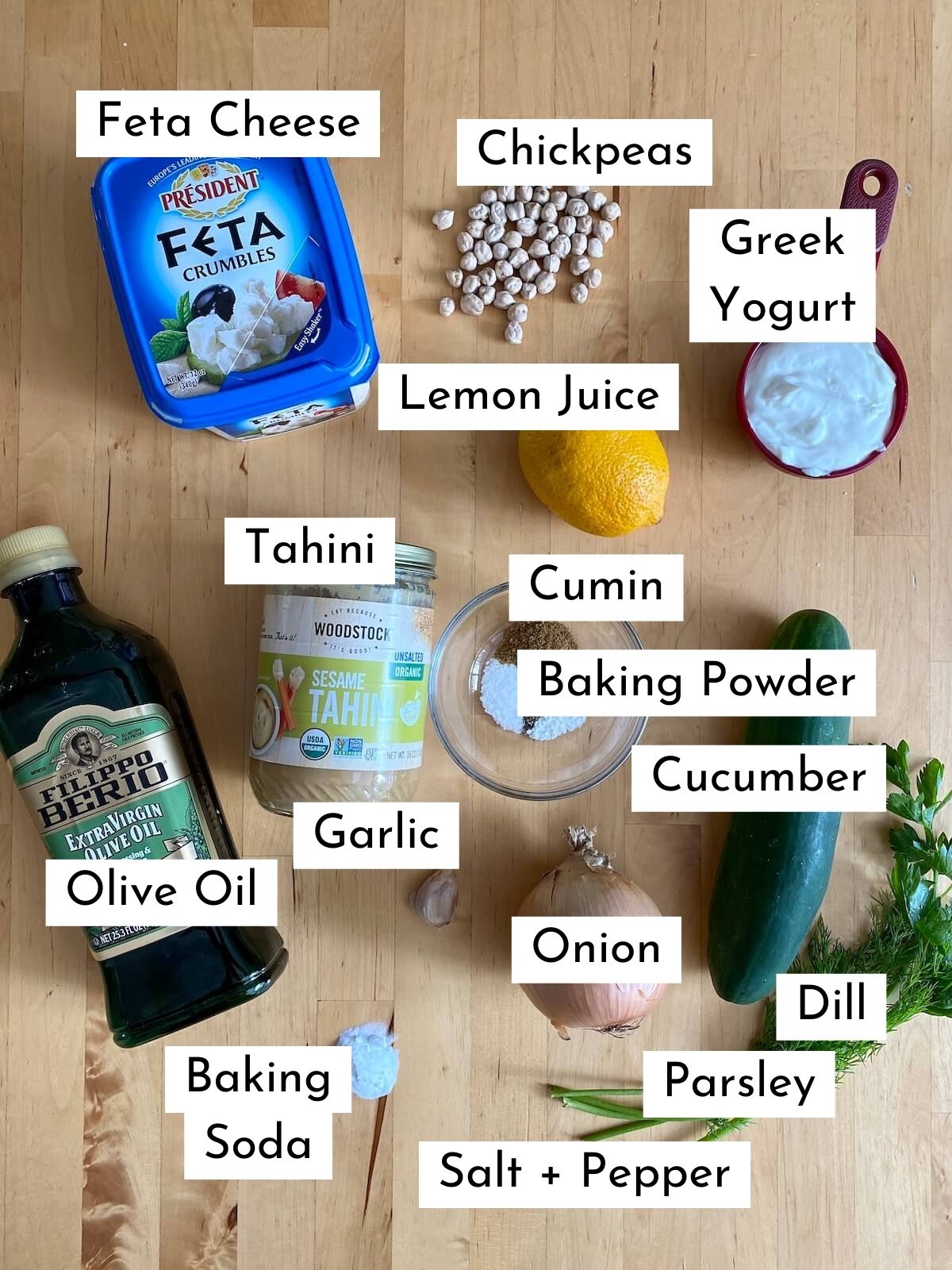 The ingredients to make falafel gyros on a wooden countertop. The ingredients are labeled with text stating what each one is. They include feta cheese, chickpeas, Greek yogurt, lemon juice, tahini, cumin, baking powder, olive oil, garlic, onion, cucumber, dill, parsley, baking soda, salt, and pepper.