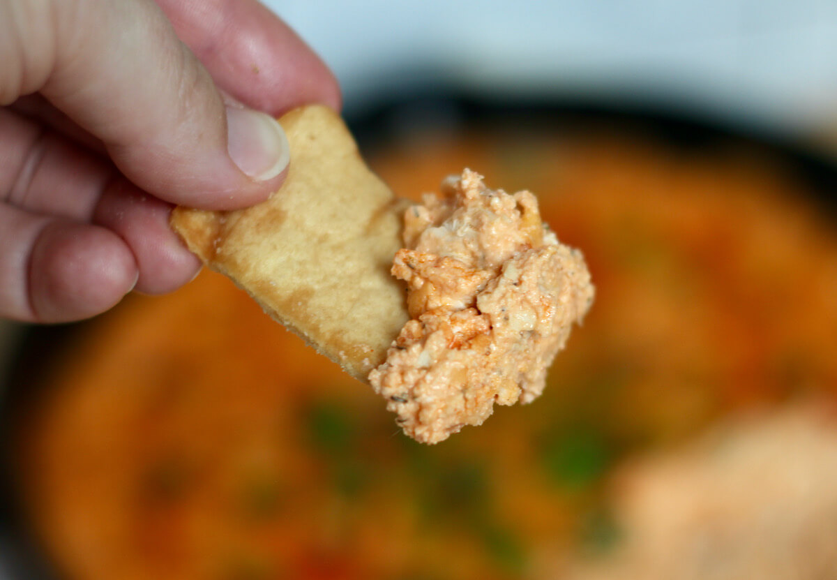 A pita chip being held above a skillet filled with hot wing dip. There is some dip on the pita chip..