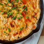 A skillet filled with Greek yogurt buffalo chicken dip garnished with green onions and hot sauce.