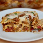 A piece of lasagna al forno on a small white plate. The tray of lasagna is out of focus in the background.