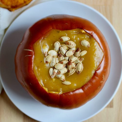 A roasted pumpkin bowl filled with pumpkin soup. The pumpkin bowl is sitting on a small white plate and the soup is topped with roasted pumpkin seeds.